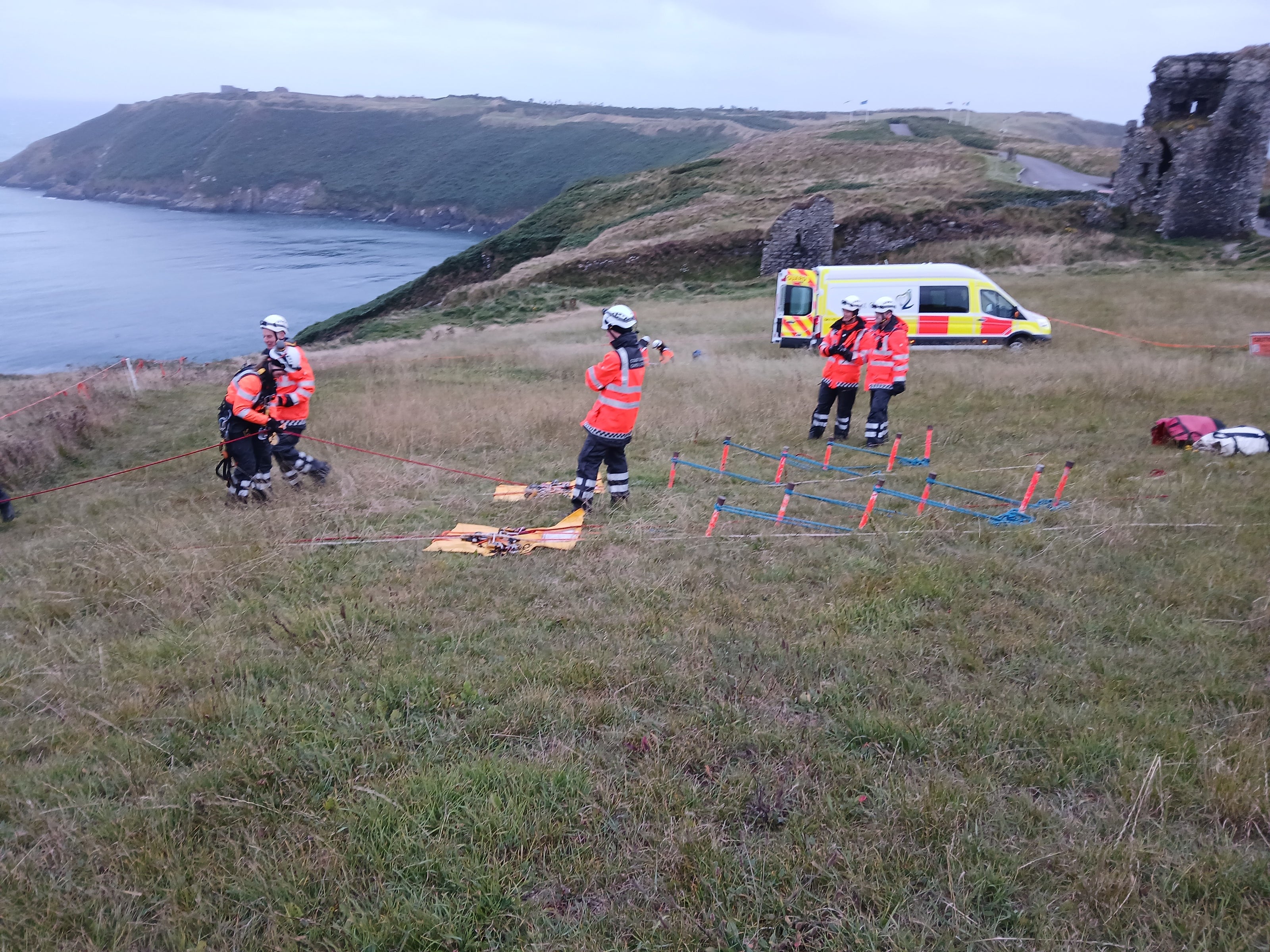 Emergency responders in high-visibility orange uniforms and helmets conduct an outdoor assessment on a grassy cliffside near the sea, with a yellow and white ambulance parked nearby.